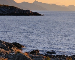 Fotografia zachytáva skoré ráno a pobrežie na Lofotoch. Do diaľky sa tiahnu horské chrbty zvažujúce sa do vôd Atlantiku.
